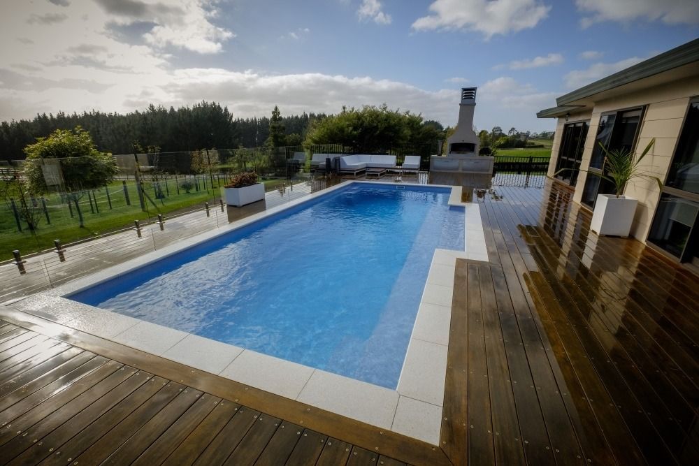 Rectangular outdoor pool with blue water, wooden deck, and landscaped garden under a cloudy sky