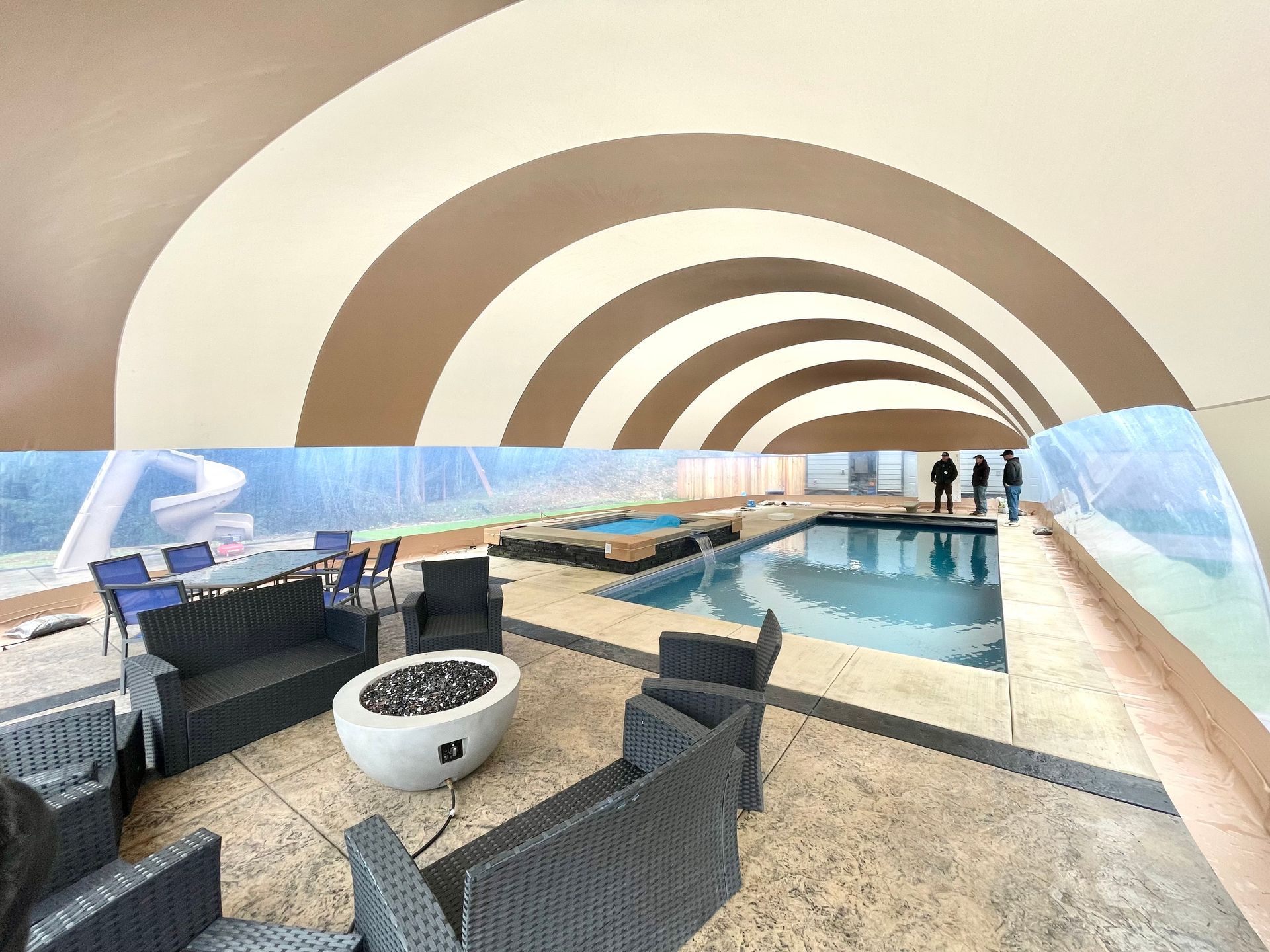Curved rooftop pool with wicker lounge chairs, a fire pit, and ocean view under a striped canopy