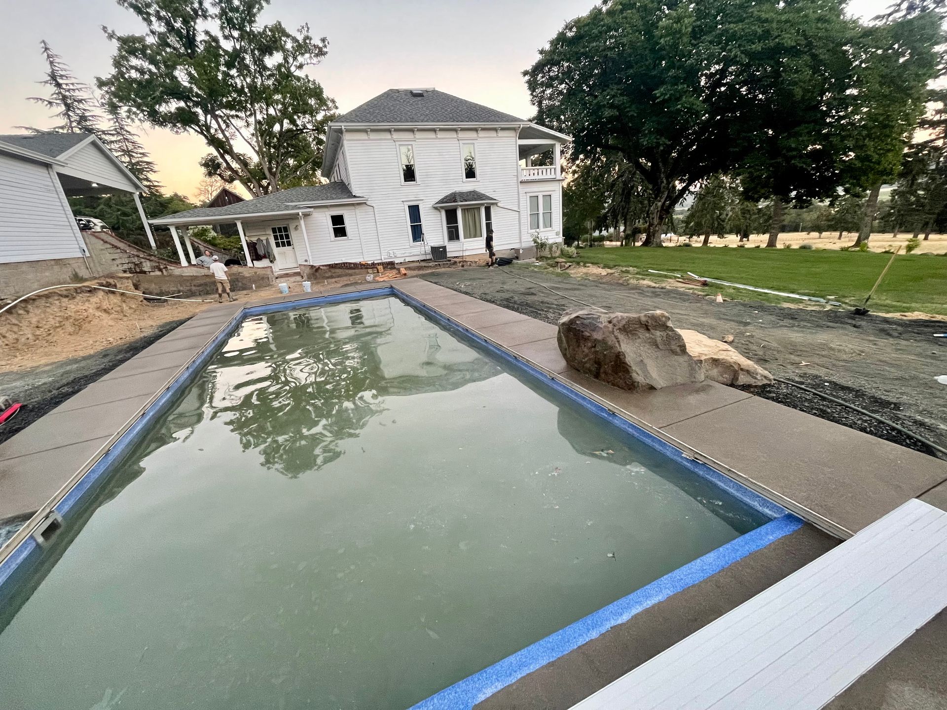 Backyard pool under construction beside a white house, with unfinished landscaping and a covered walkway