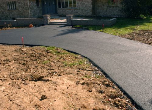 Freshly paved curved driveway leading to a house entrance, bordered by dirt and grass with survey flags.