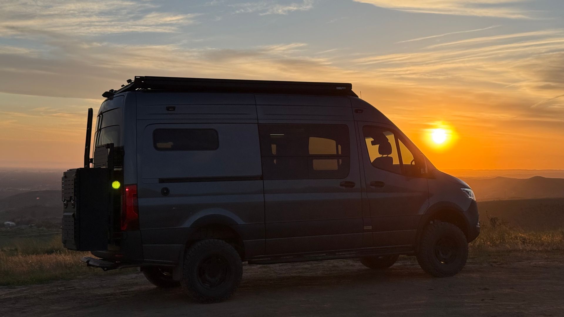 Dark van parked on a dirt road at sunset, silhouetted against a golden sky.