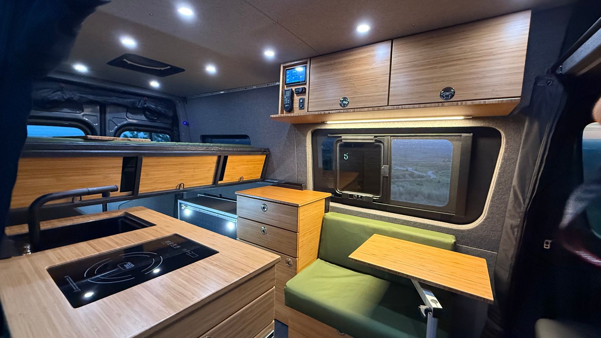 Compact van interior with wood cabinetry, benches, a table, and a skylight-lit workspace in warm lighting