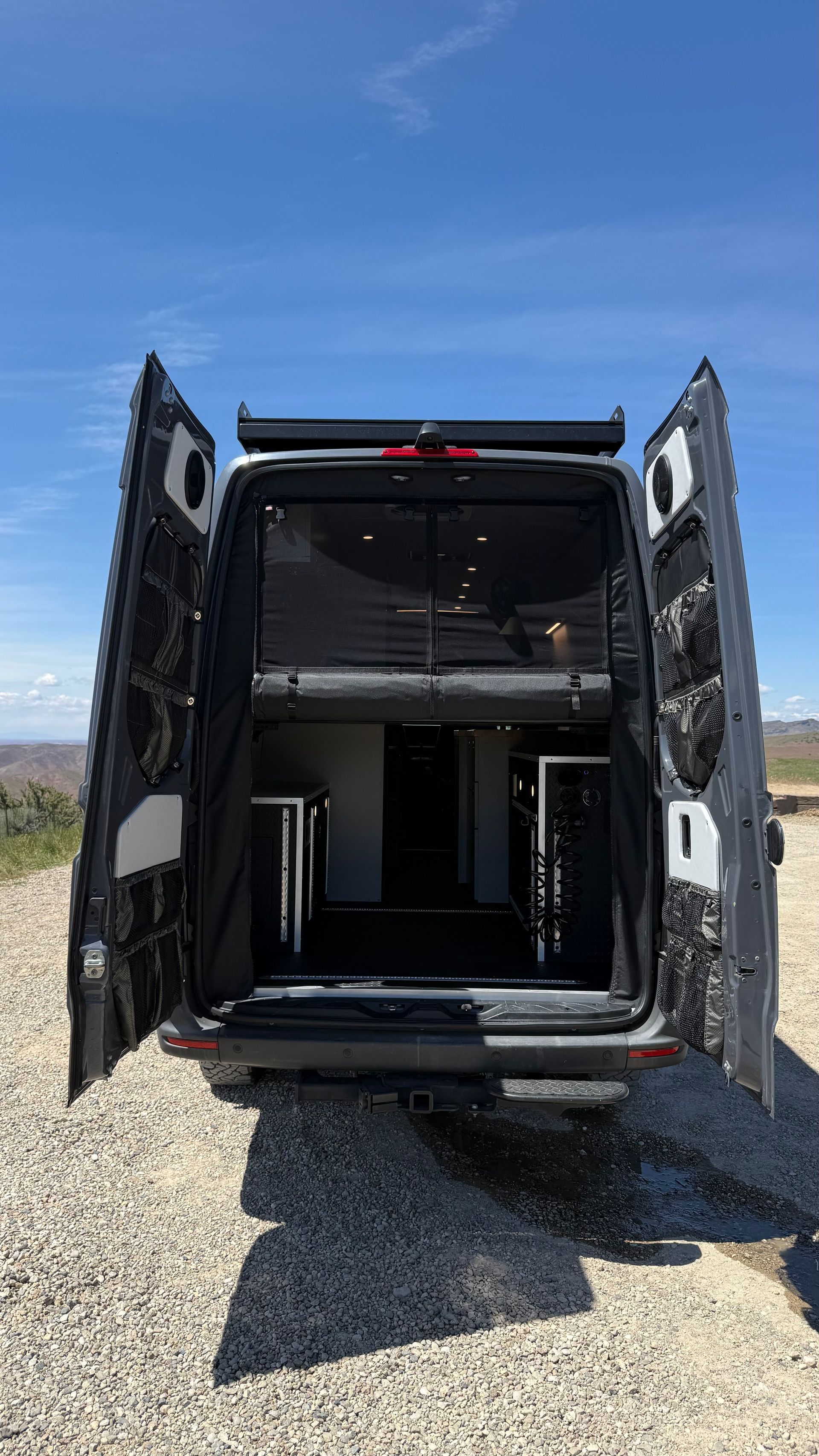 Open rear doors of a black van with a metal lift platform inside, parked on gravel under a blue sky