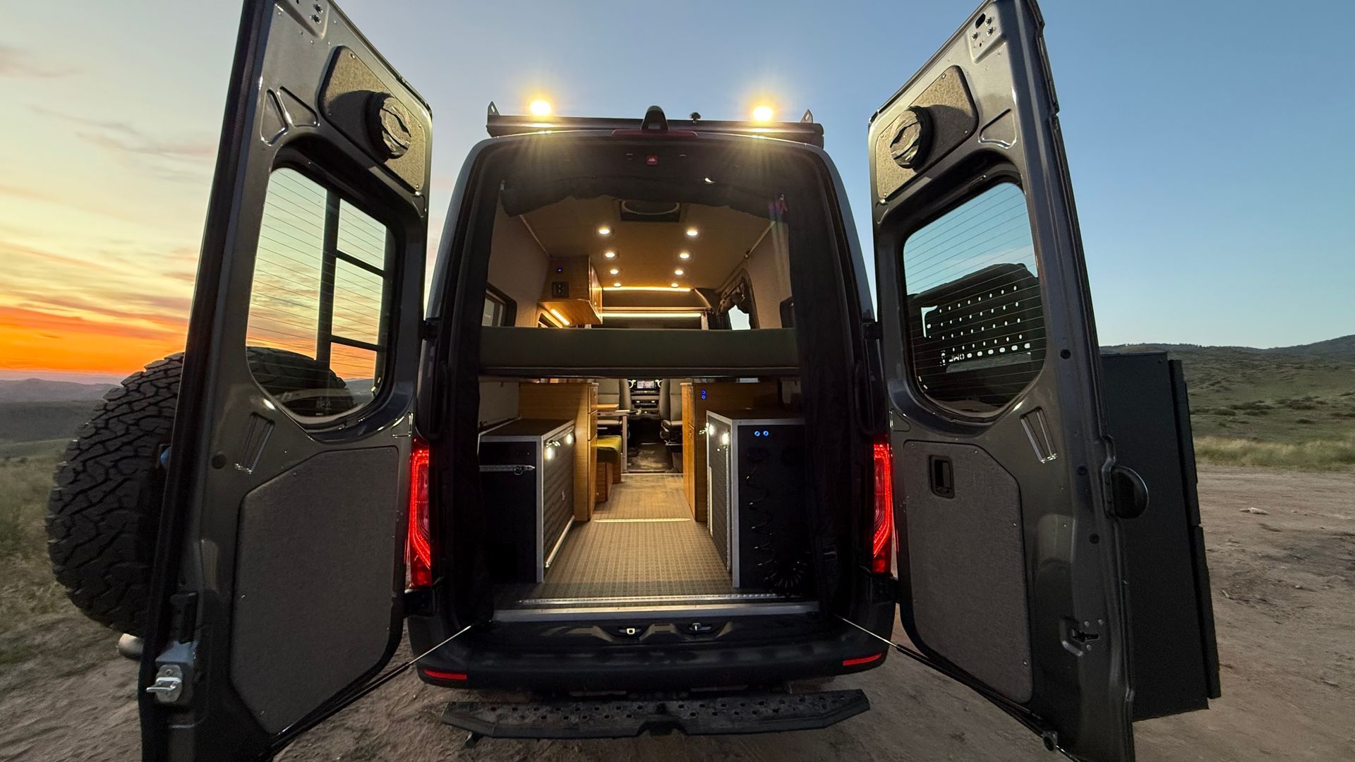 Open van with rear doors open at sunset, showing a lit interior and bed inside.