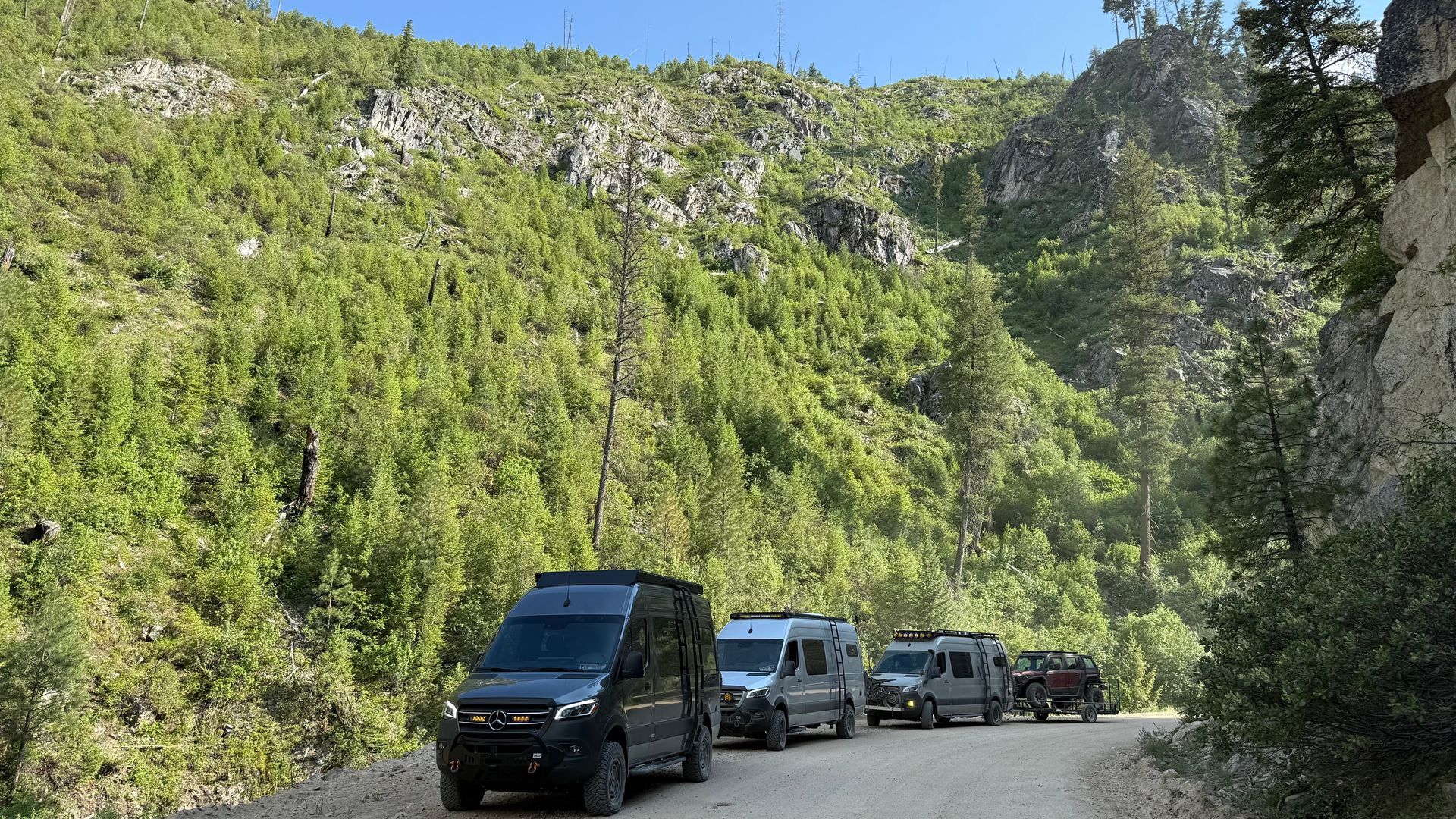 Vehicles driving on a narrow mountain road beside a steep green cliff.