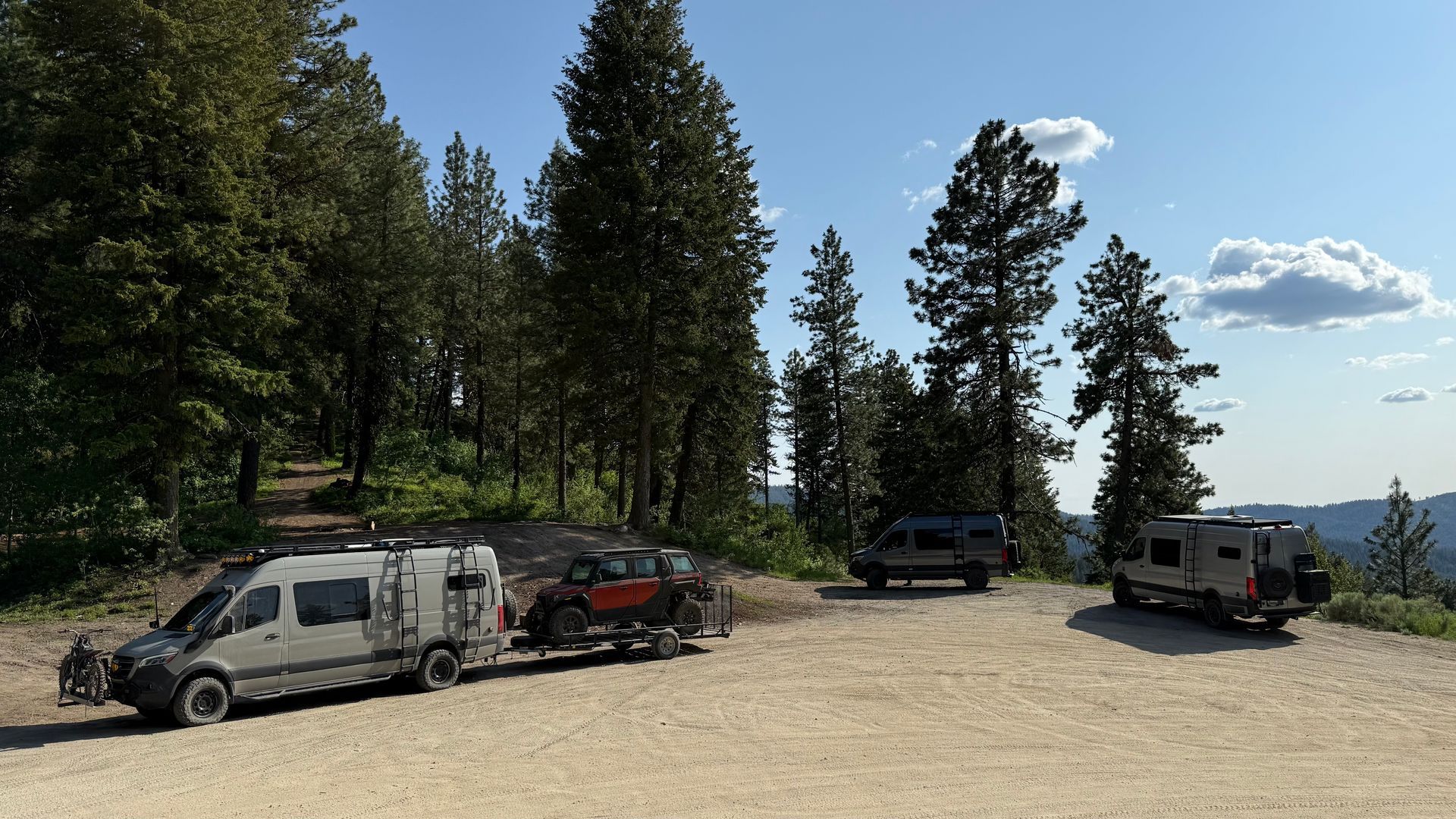 Several parked vehicles in a forest clearing under a blue sky, including a white van and off-road trucks.