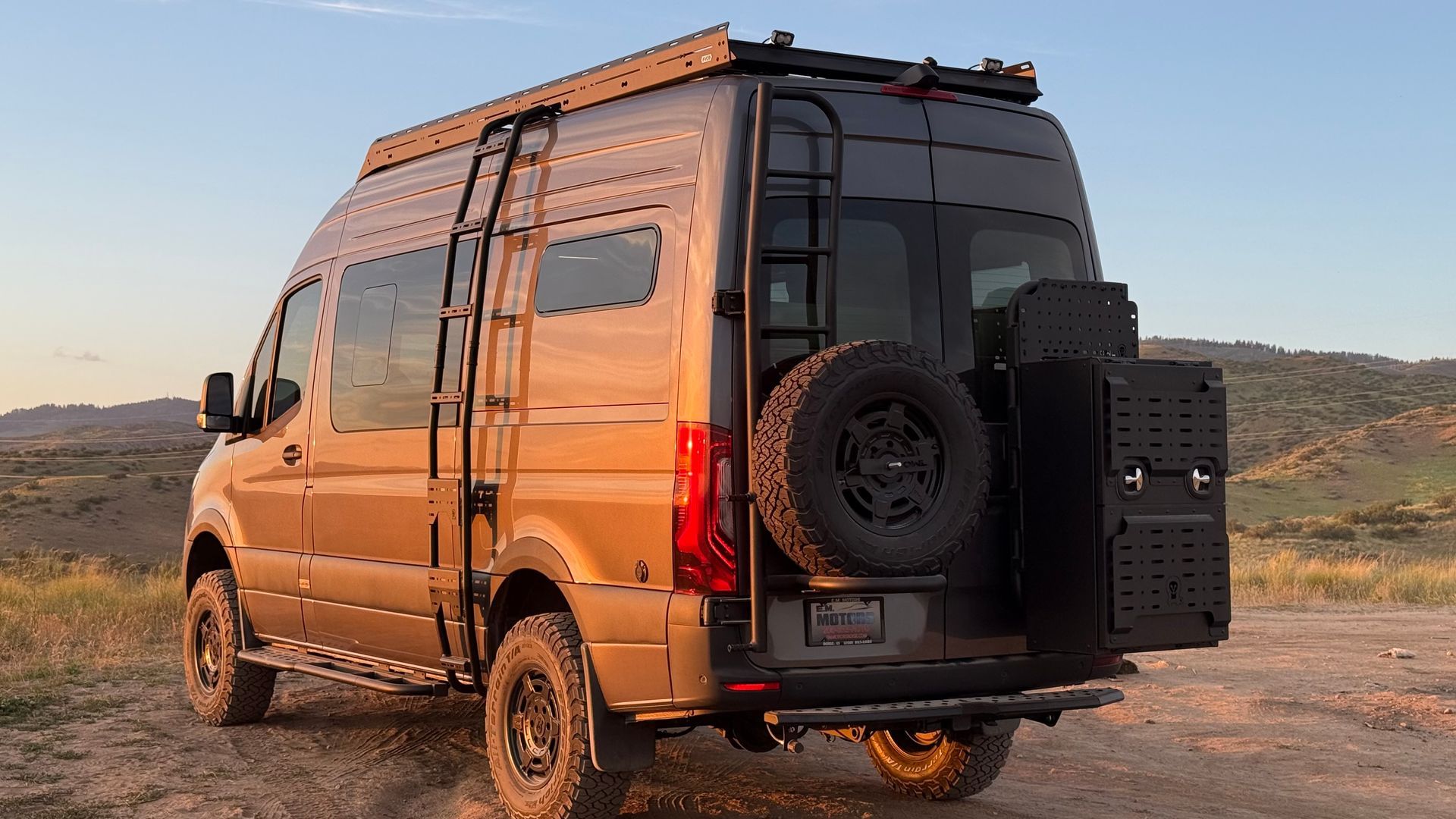 Bronze camper van on a dirt overlook at sunset, rear view with spare tire and gear box.