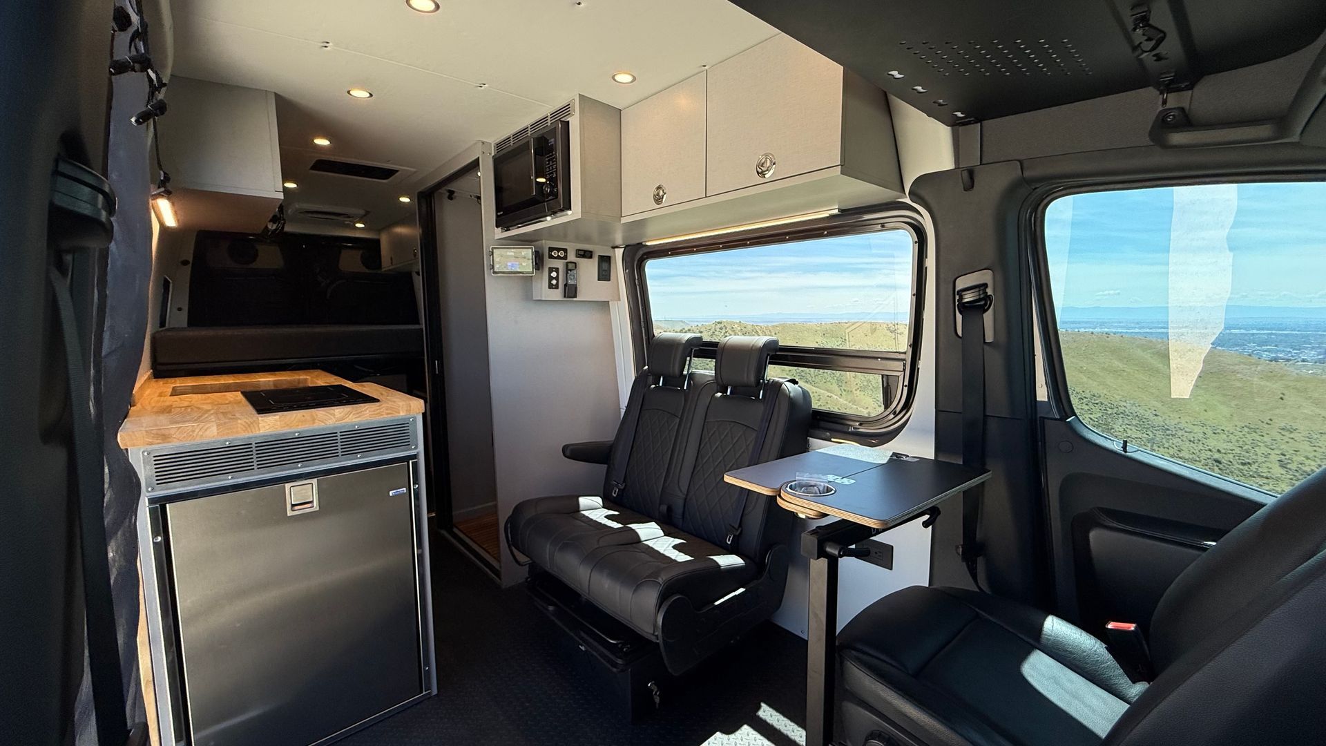 Inside a camper van, a compact kitchen and two black seats face a bright window with open countryside views