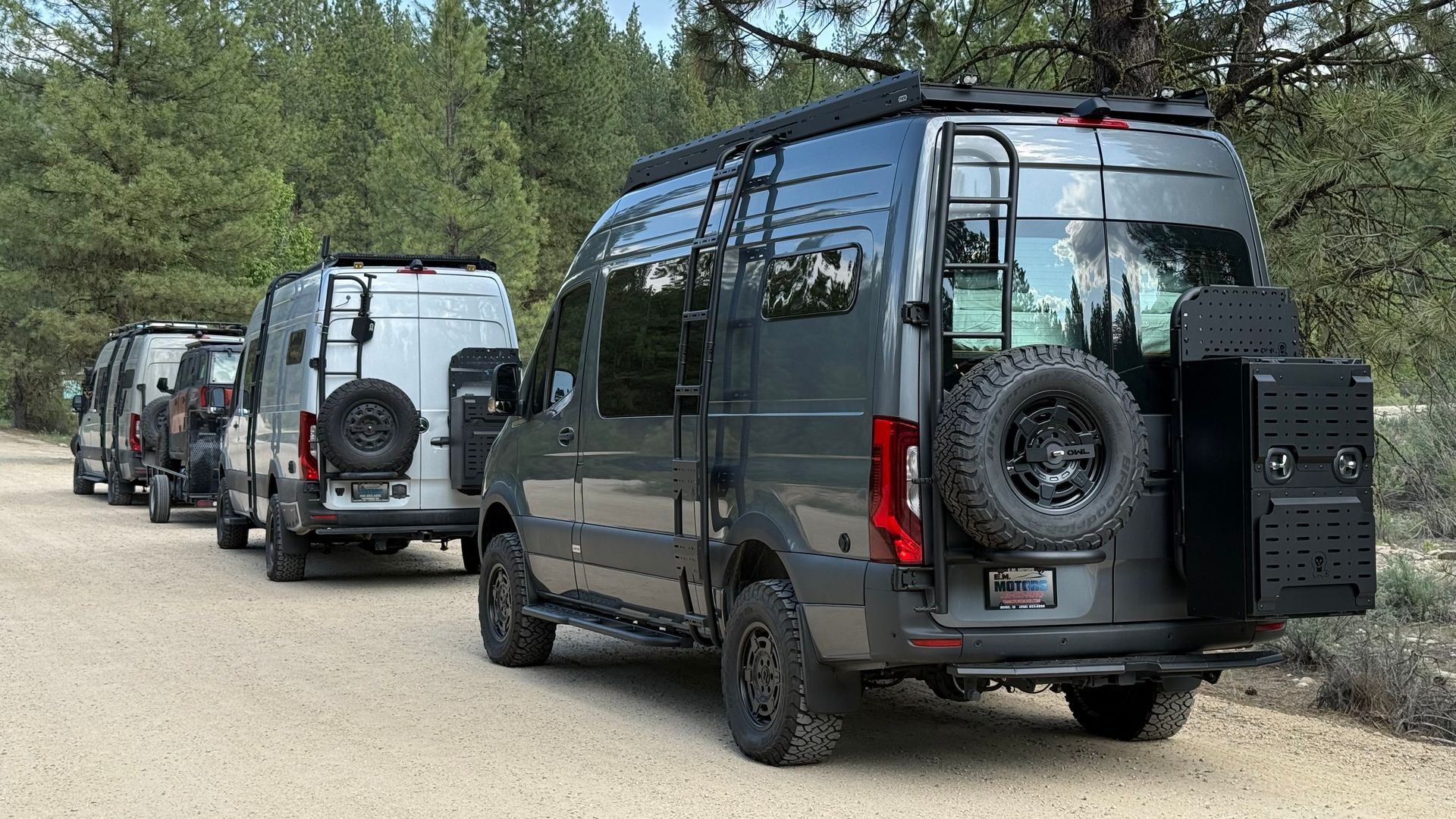 Three dark gray vans parked in a wooded dirt lot, each with a rear-mounted spare tire.