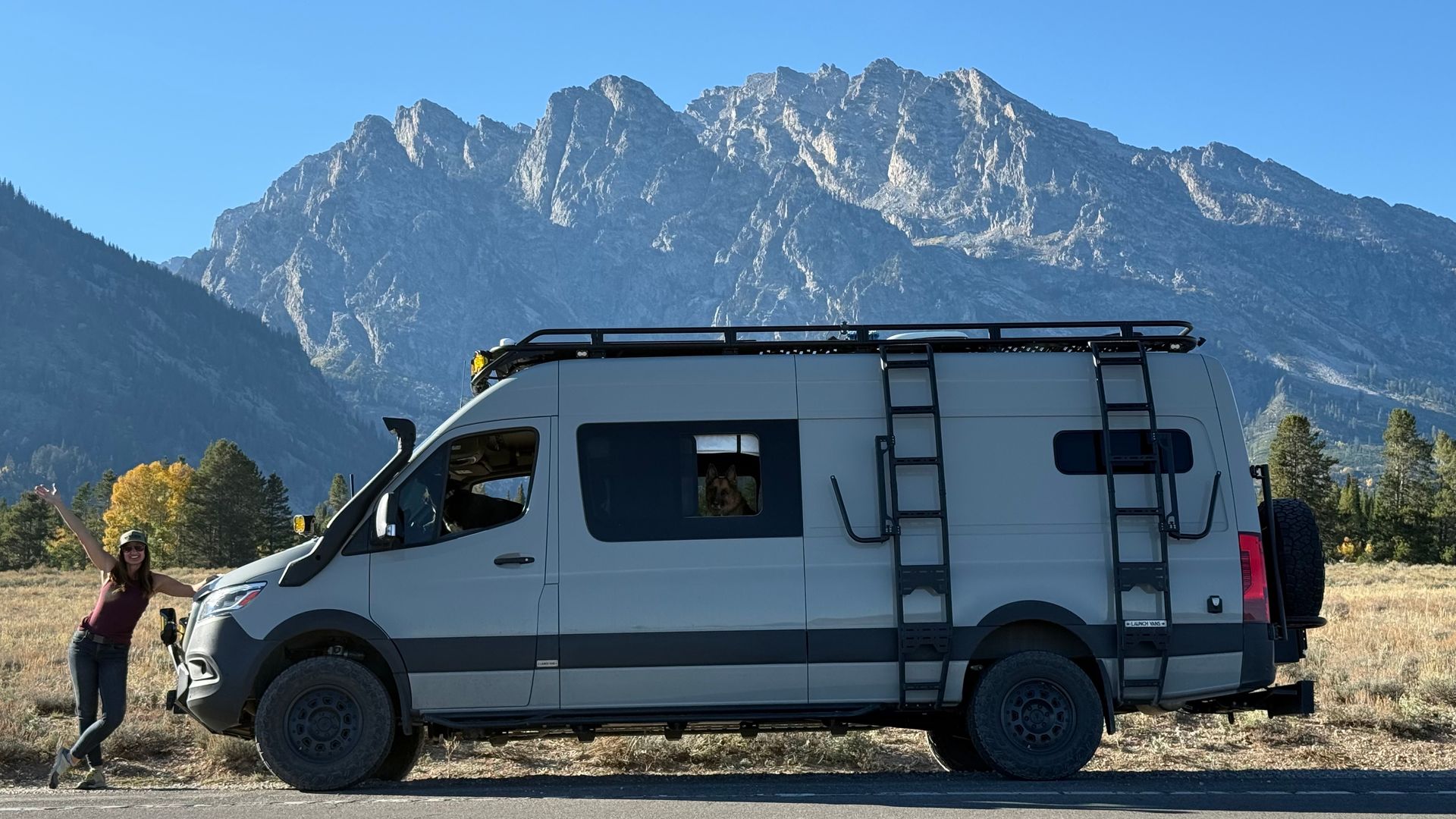 White camper van with roof rack parked in front of mountains, with a person beside it