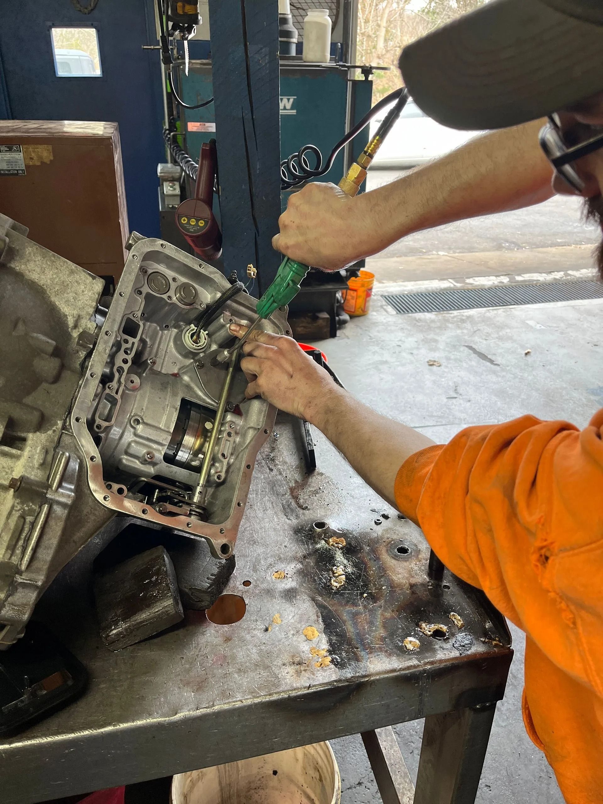 Mechanic cleaning a car transmission with a green hose in a shop.