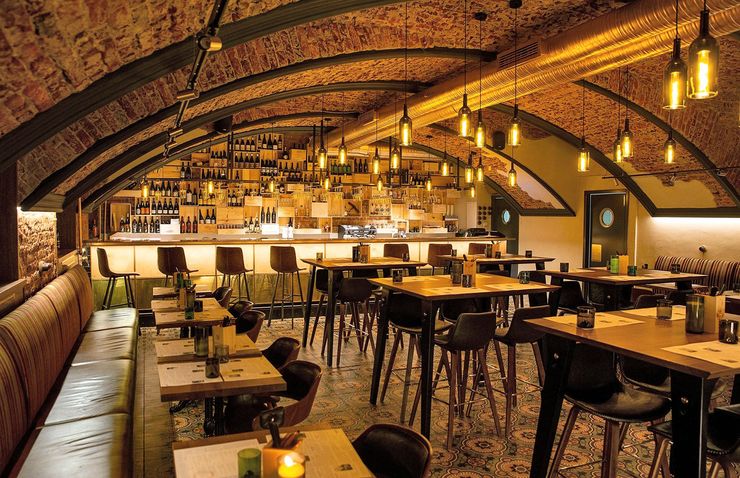 Restaurant interior with arched brick ceiling, bar, tables, and chairs; warm lighting.