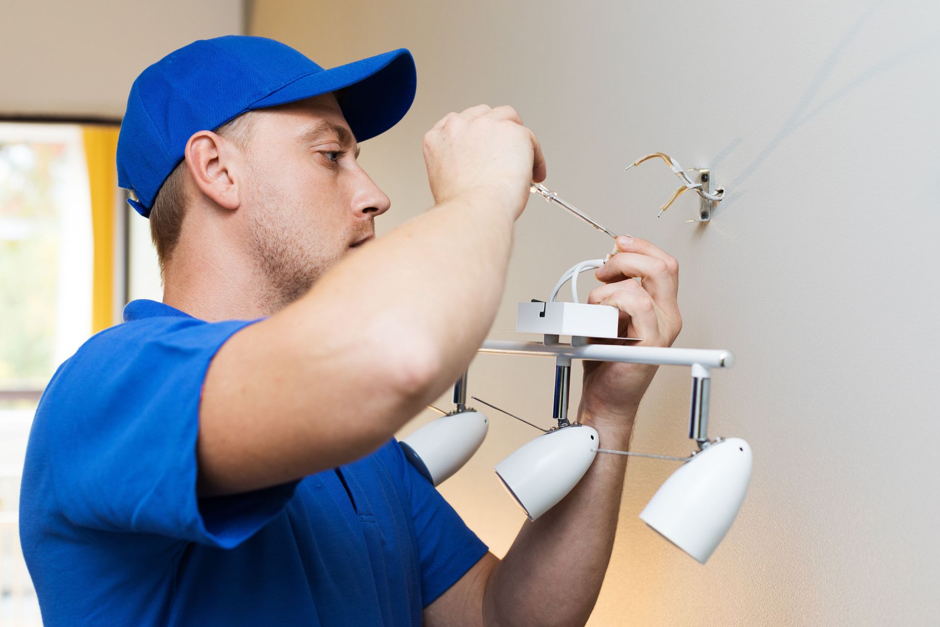 Man in blue shirt and cap installing a white light fixture on a wall.