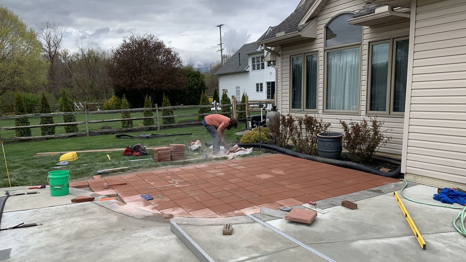 A person laying red bricks to construct a patio in a grassy backyard next to a house.