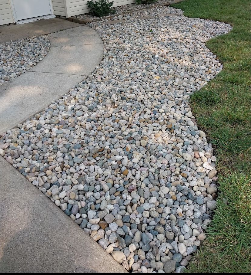 A curved concrete walkway bordered by a decorative bed of gray and tan river rocks next to a green lawn.