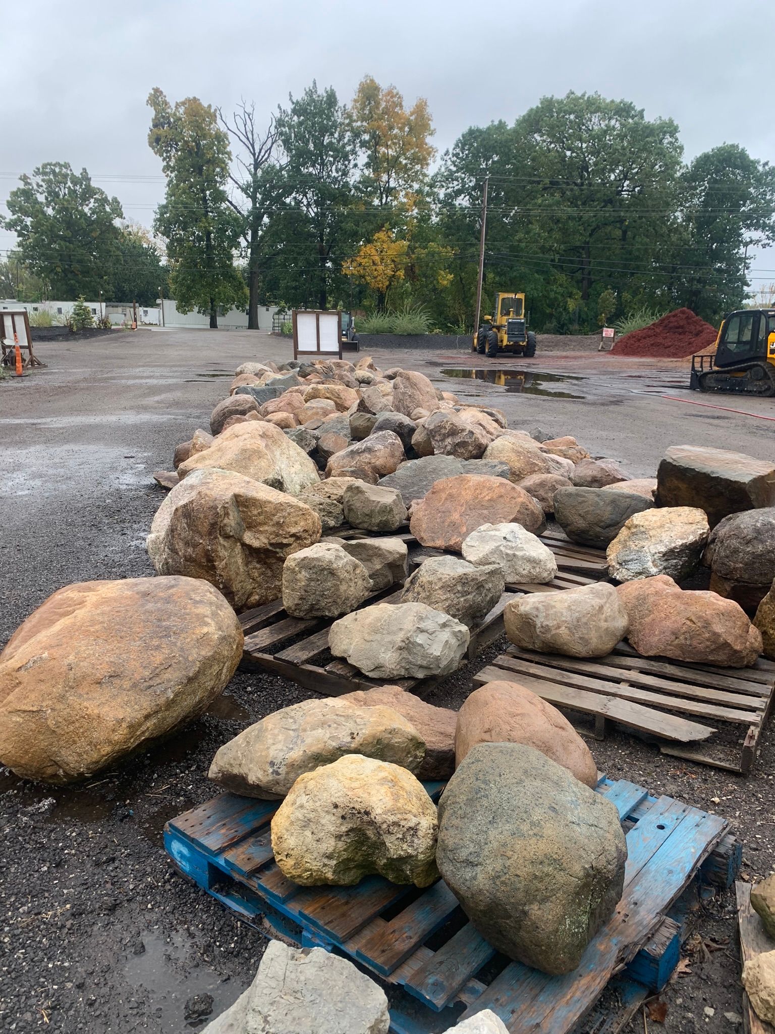 Large landscape boulders resting on wooden pallets in an outdoor supply yard with trees and heavy machinery in the distance.