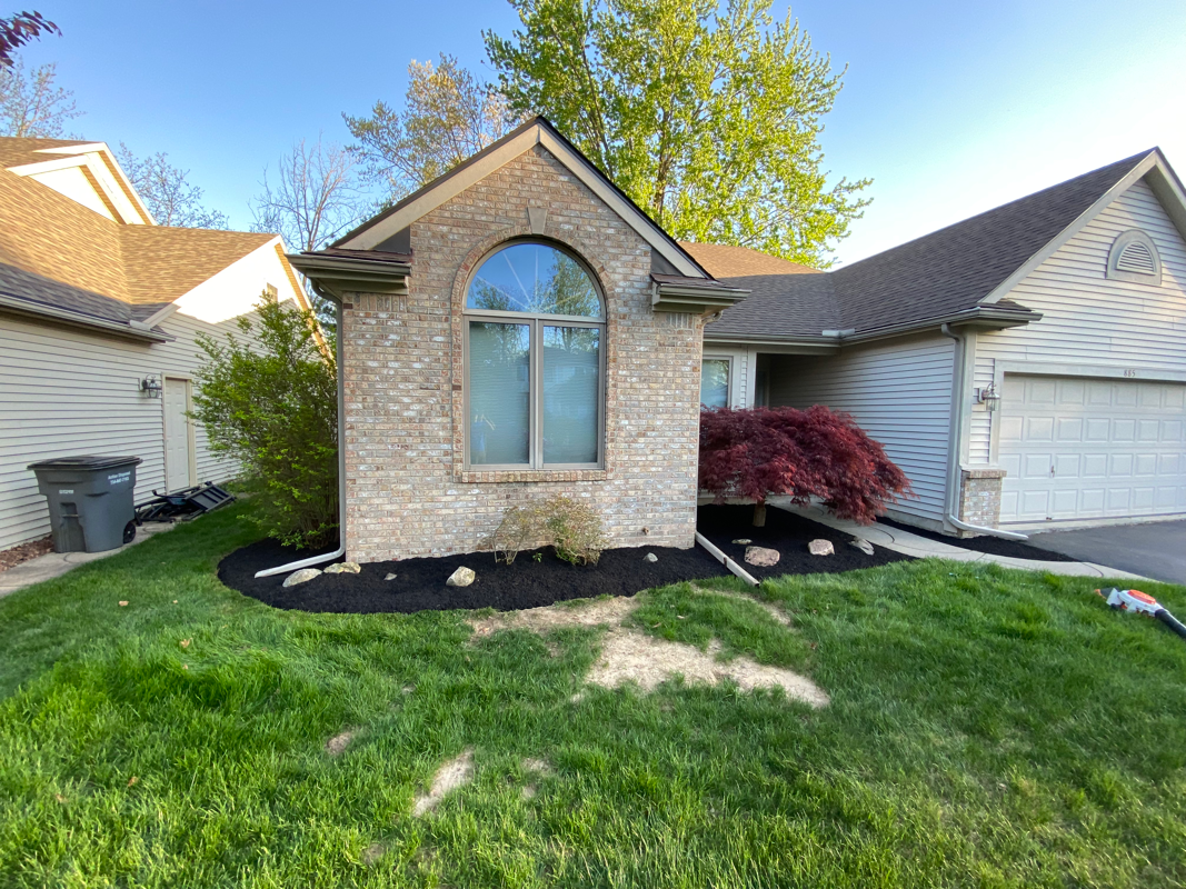 A single-story suburban house with brick facade, arched window, and a dark mulch garden bed in front of a green lawn.