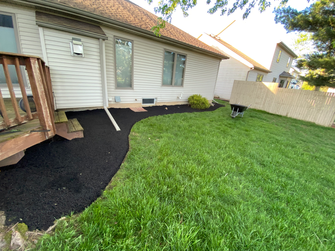 A side view of a house with light beige siding, a wooden deck, and a freshly mulched garden bed bordering a green lawn.