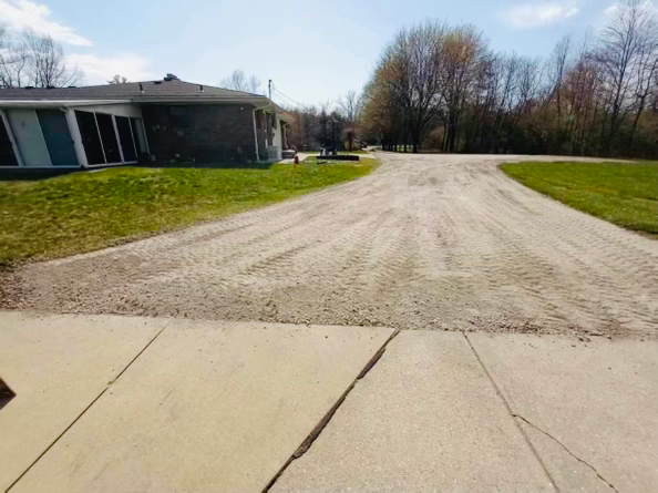 A view from a concrete driveway onto a wide gravel driveway leading toward a house and a treeline under a blue sky.
