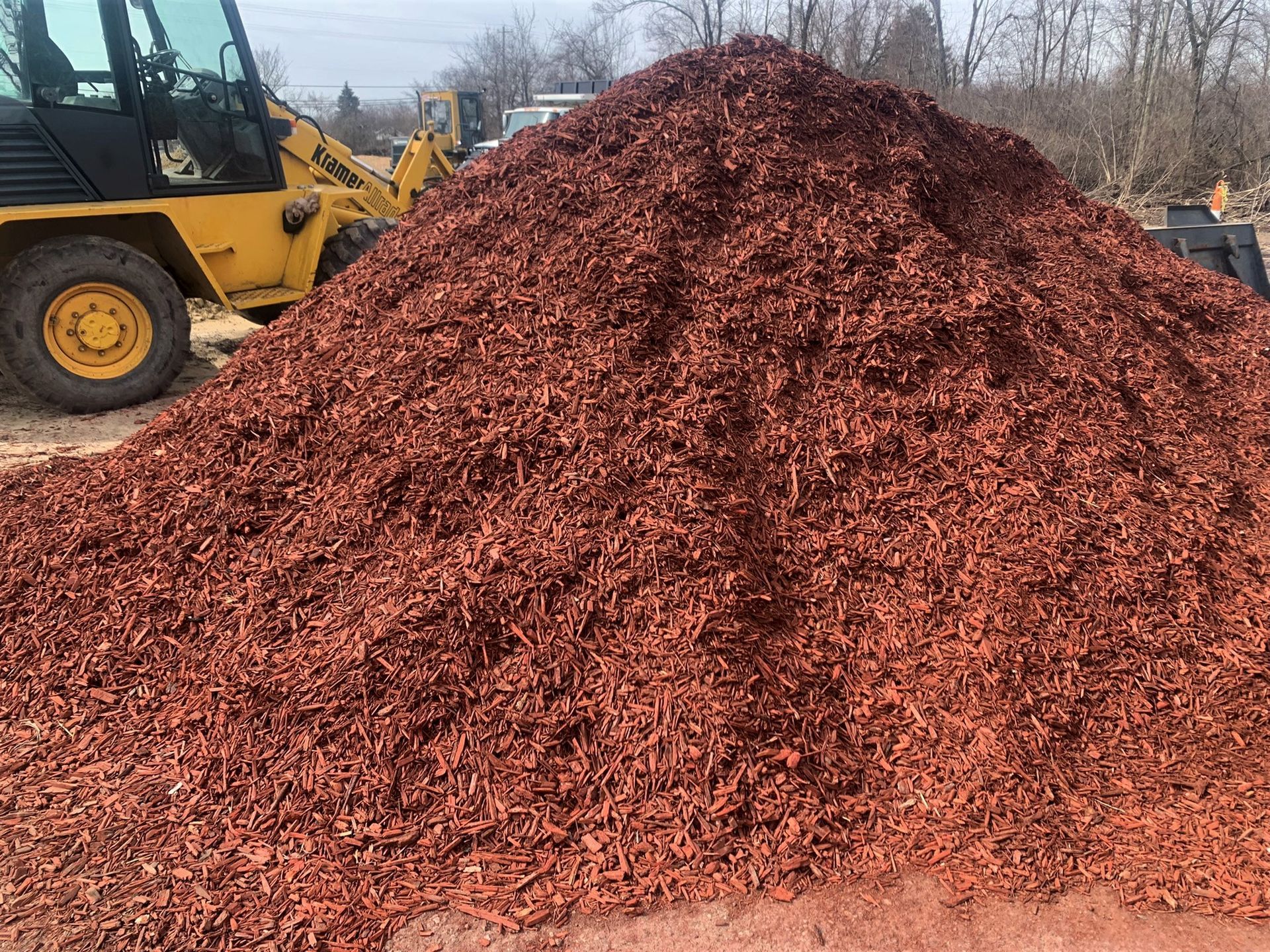 A large pile of shredded red mulch sits next to a yellow construction tractor on a dirt surface.