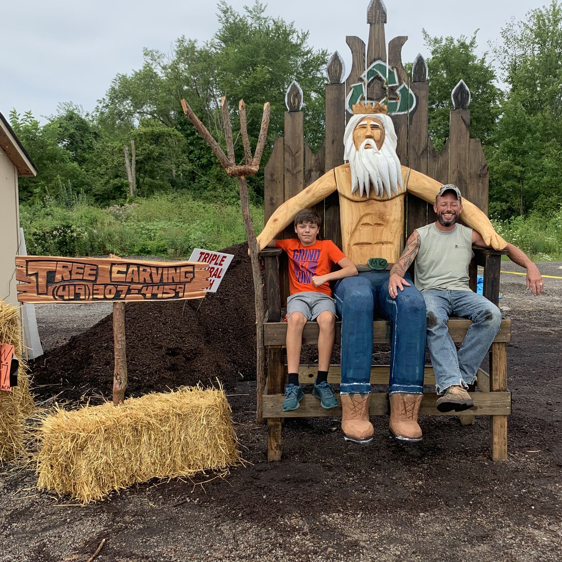 A child and an adult sit on a large, carved wooden throne depicting a figure with a beard, holding a wooden trident.
