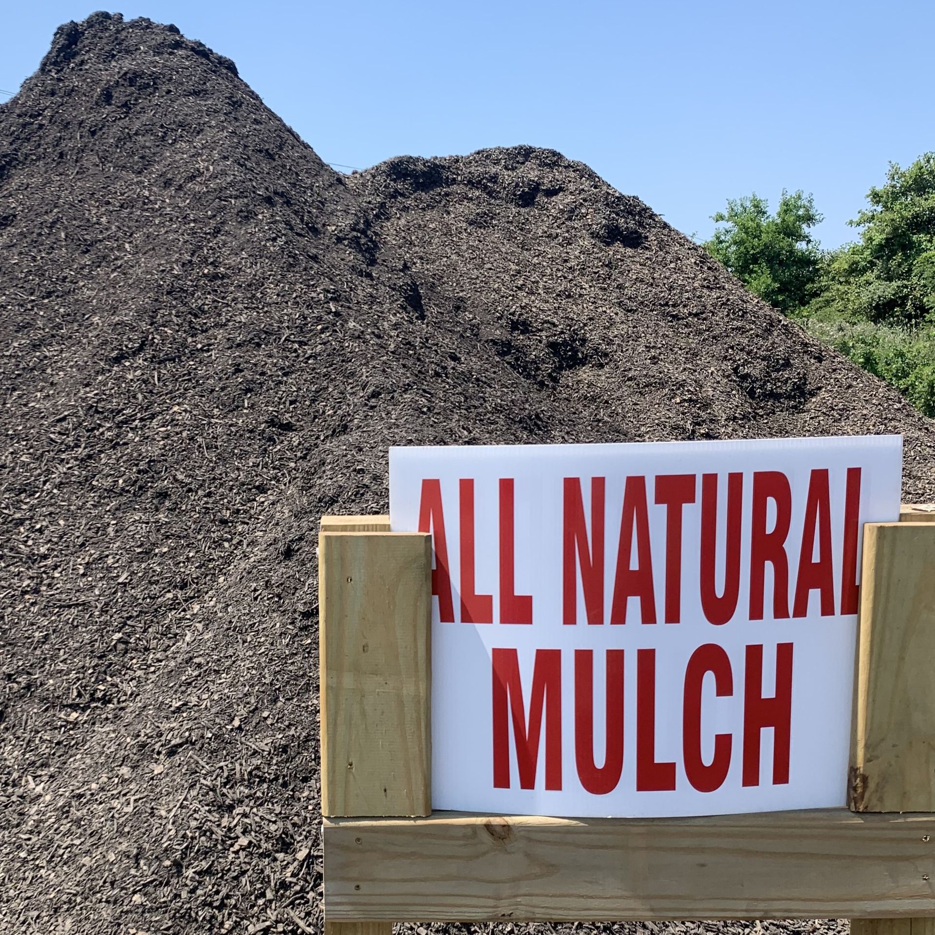 A large pile of dark, natural wood mulch behind a wooden sign that reads 