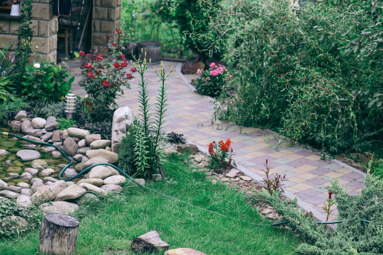 A stone pathway winds through a lush garden with a small pond, blooming flowers, and green shrubbery.