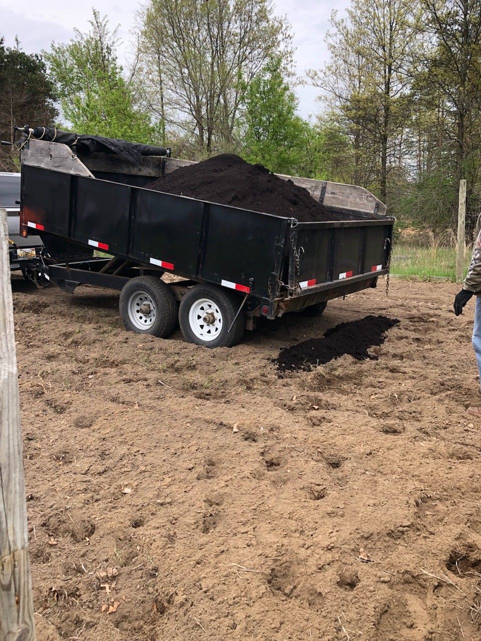 A black dump trailer filled with dark soil sits in a dirt field next to a vehicle, with a person standing nearby.