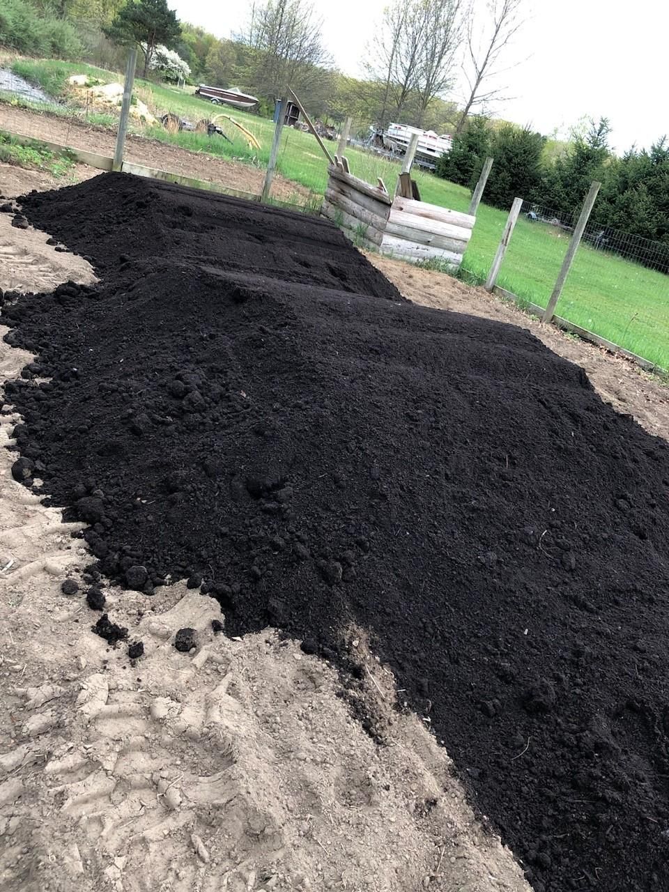 Large piles of dark, rich organic soil or compost sit on a bare dirt field near a wooden fence and grassy area.