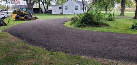 A newly installed dark gravel driveway curves through a green lawn toward a small white house, with a yellow skid steer.