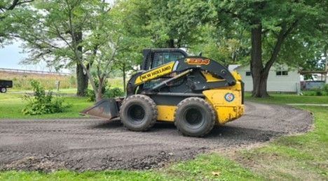 A yellow New Holland skid steer loader sits on a gravel driveway in a grassy, tree-lined residential yard.