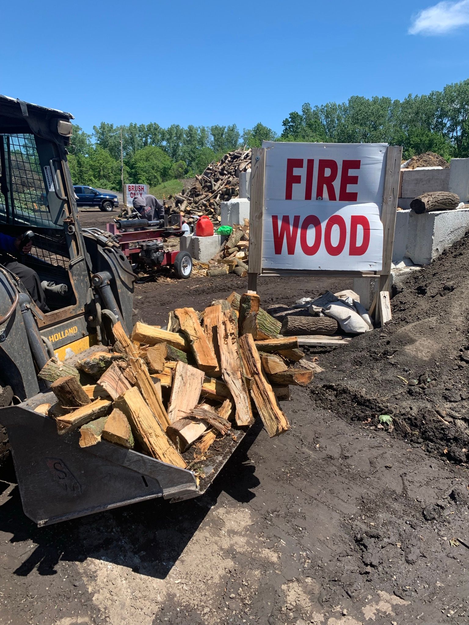 A skid steer loader filled with split firewood sits next to a white sign with red text that reads 