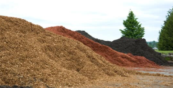 Three large piles of garden mulch in shades of brown, red, and dark charcoal sit outside near some trees.