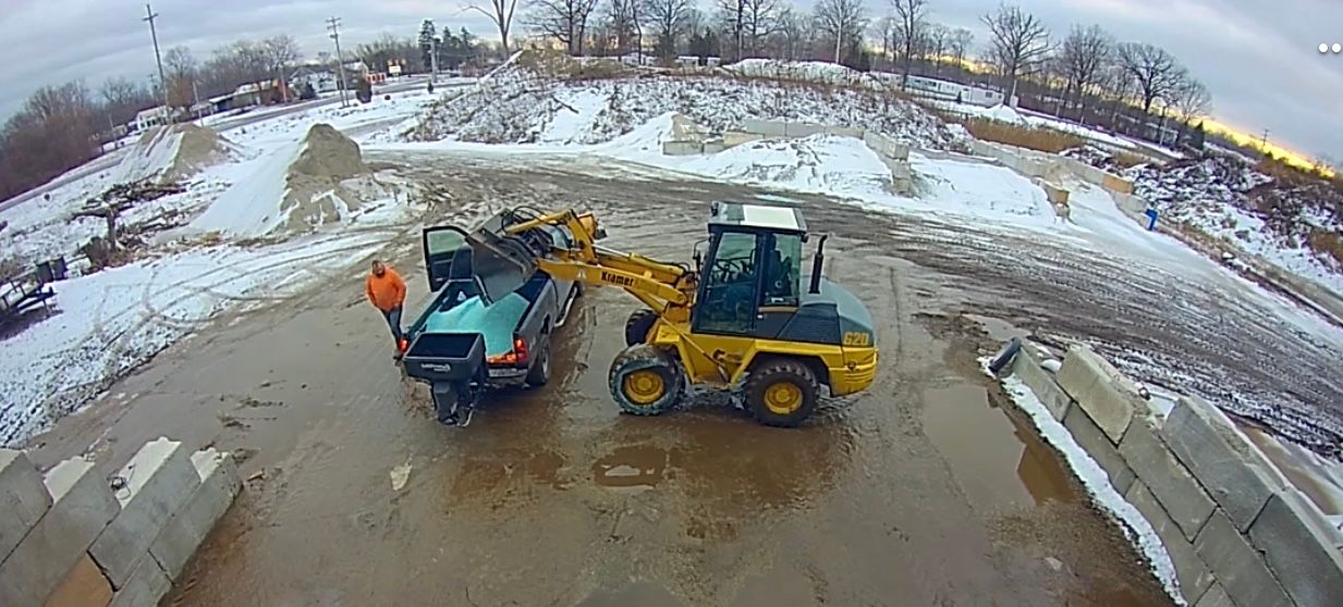 A yellow front-end loader pours material into the bed of a truck in a snowy, industrial outdoor lot.