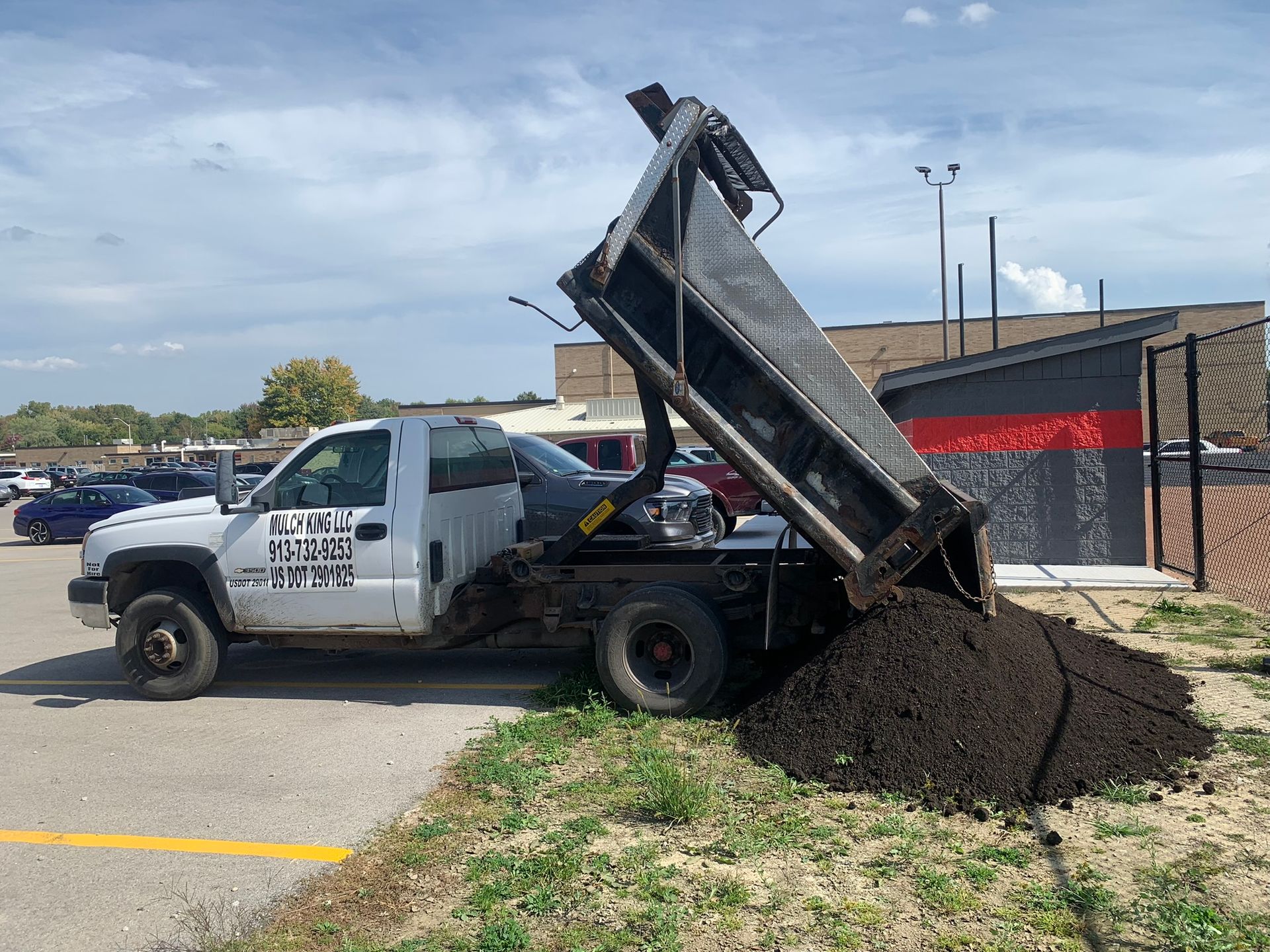 A white dump truck parked on a paved lot in the process of unloading a large pile of black mulch onto the grass.