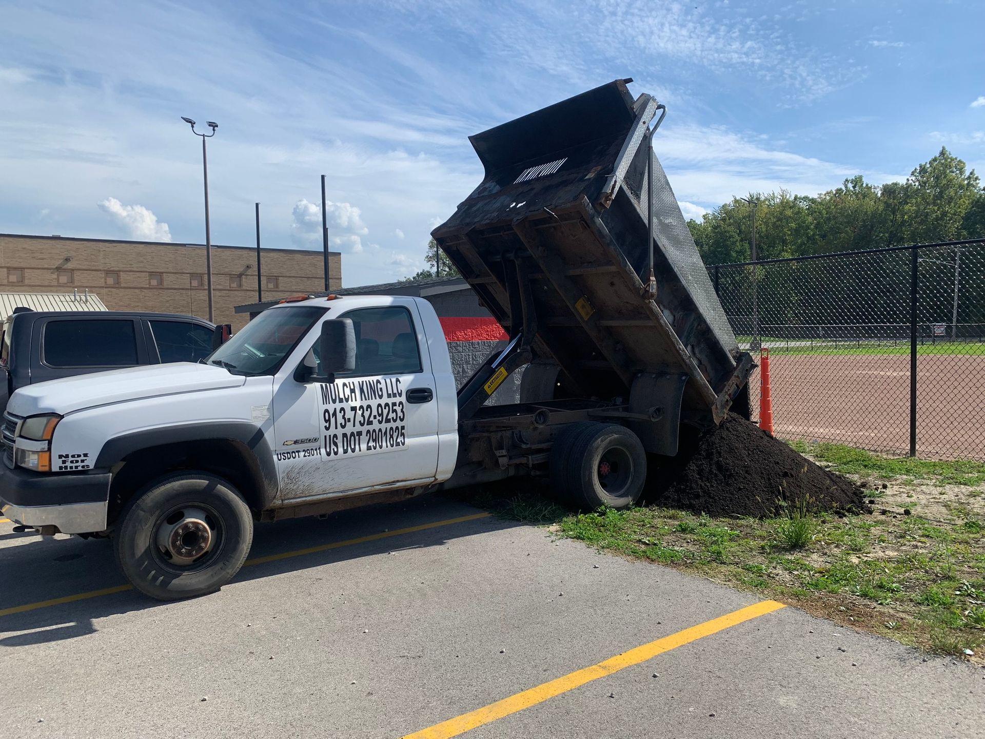 A white dump truck parked on asphalt with its bed raised, emptying a pile of dark dirt onto the ground.
