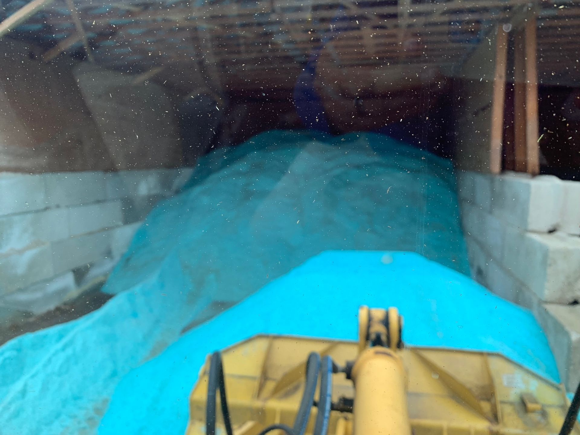A yellow loader bucket pushes a large pile of bright blue industrial material inside a storage bay with concrete walls.