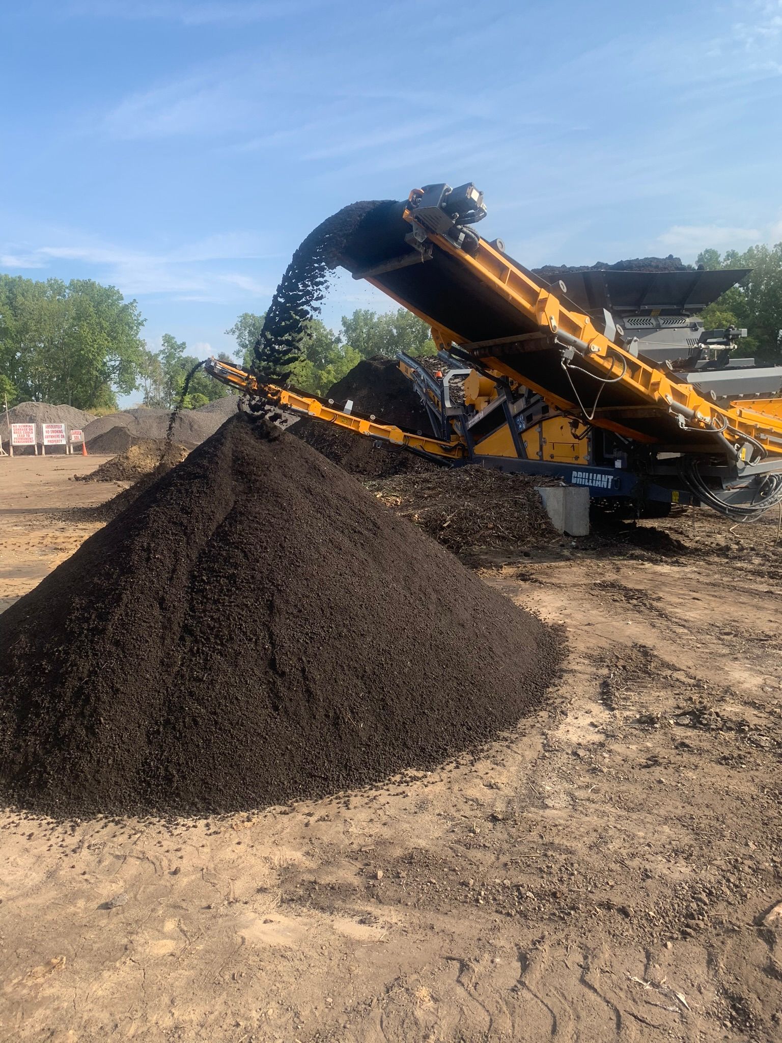 A large yellow industrial conveyor machine depositing a pile of dark, processed organic mulch outdoors under a blue sky.
