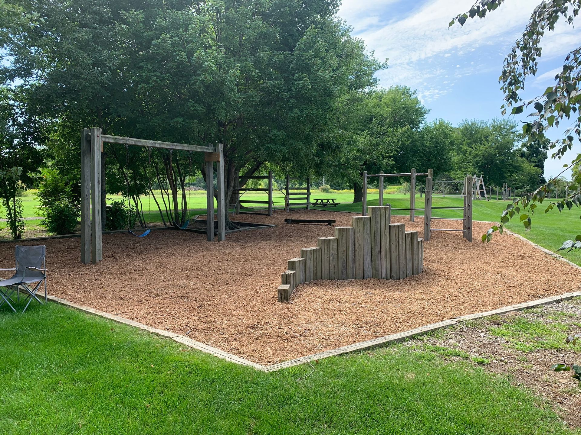 A wooden playground with monkey bars and step stumps on wood chips under trees in a grassy field.