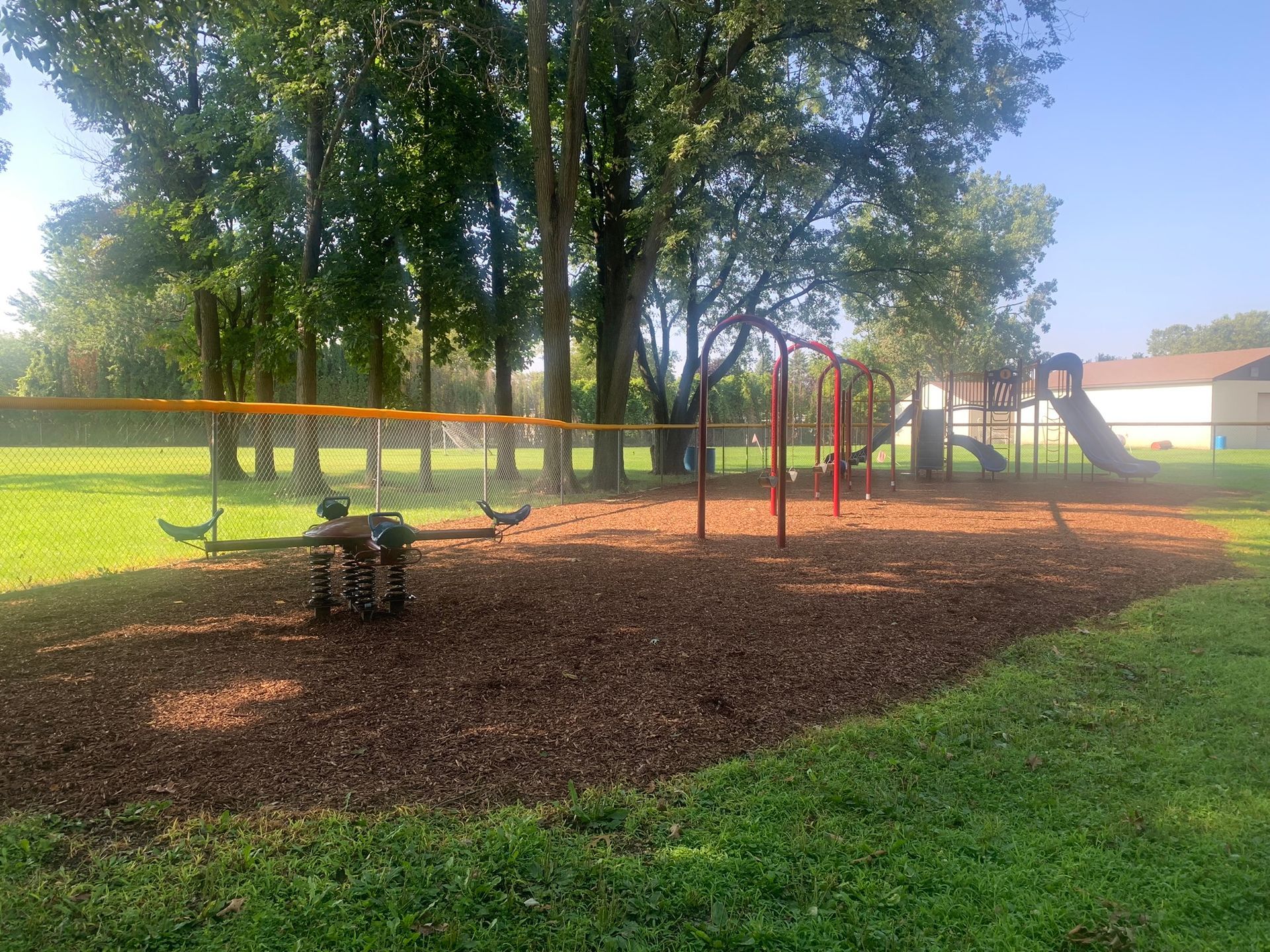 A playground with a spring rider, swings, and a slide set against a backdrop of trees on a sunny day.