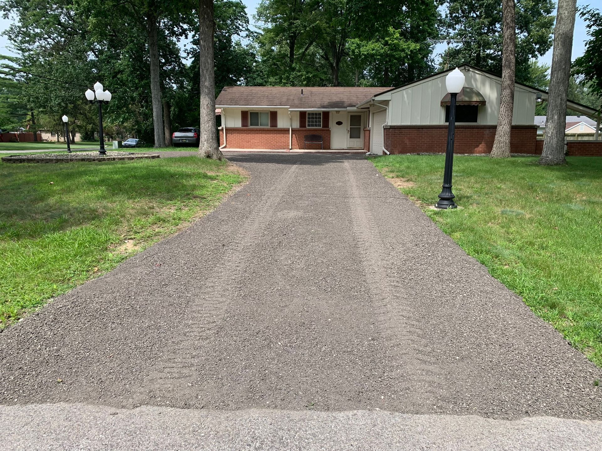 A gravel driveway leads to a single-story house with a brick facade, framed by trees and two ornate street lamps.