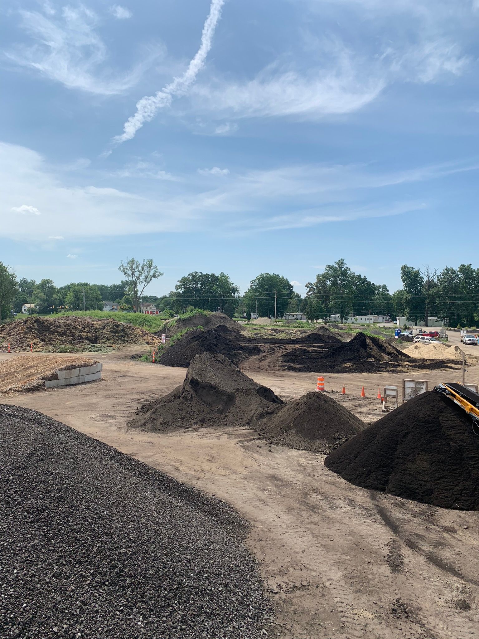 Large piles of dark soil and gravel at an outdoor construction site under a bright, partly cloudy sky.