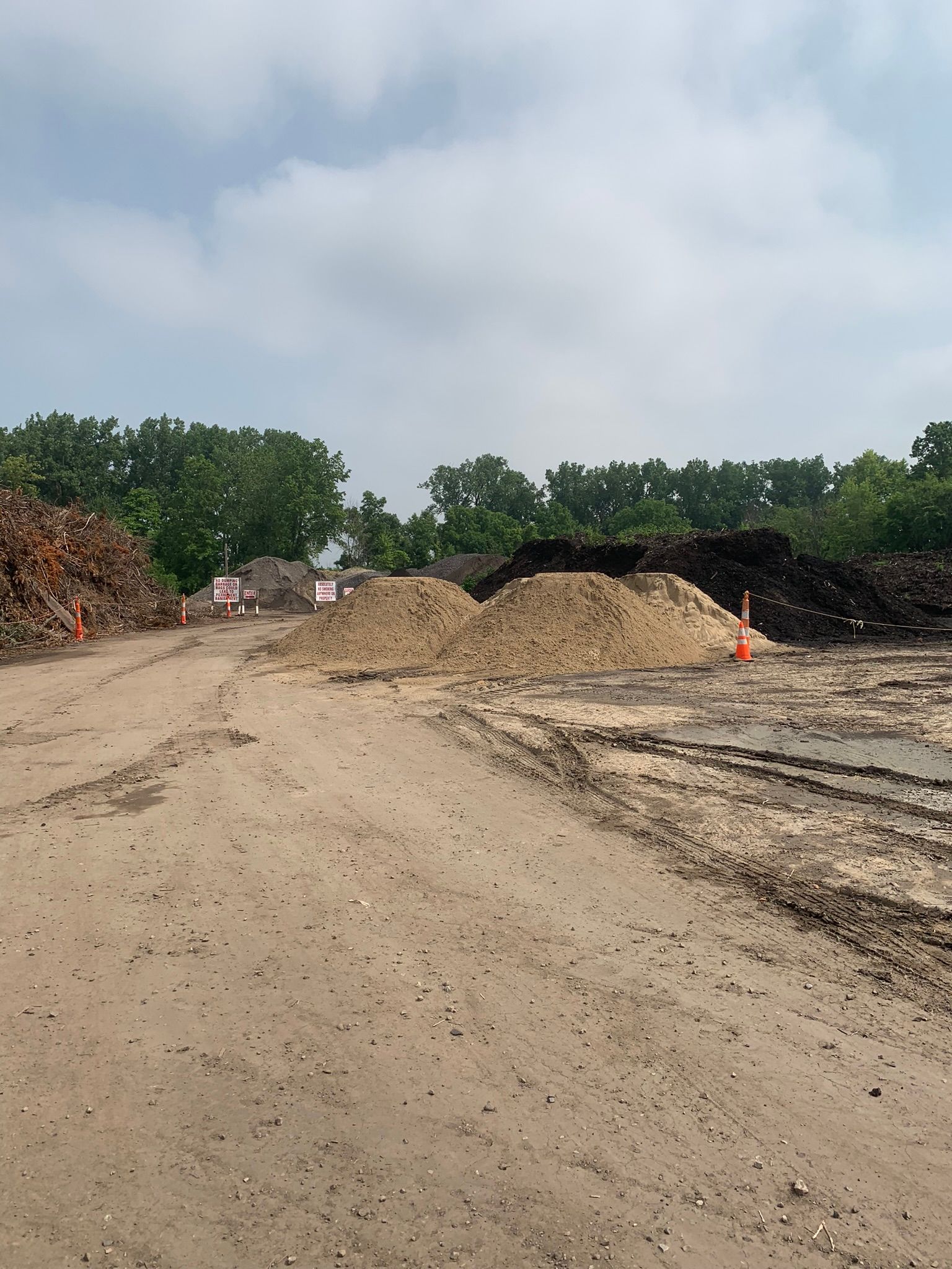 Large piles of sand and dirt sit in a dirt lot under a cloudy sky, with a treeline in the background.