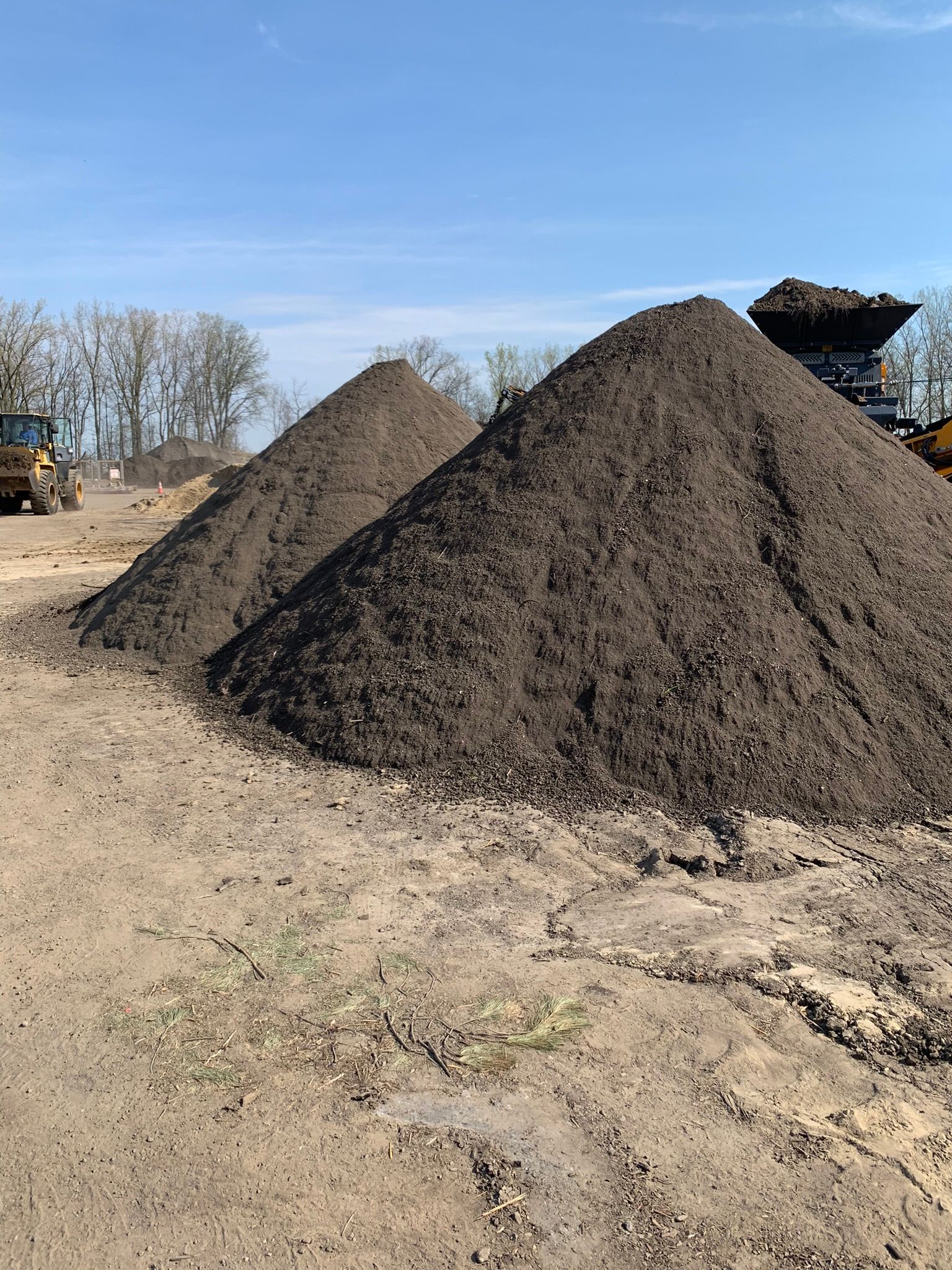 Two large, conical piles of dark soil sit on a dirt lot under a clear blue sky, with construction equipment nearby.