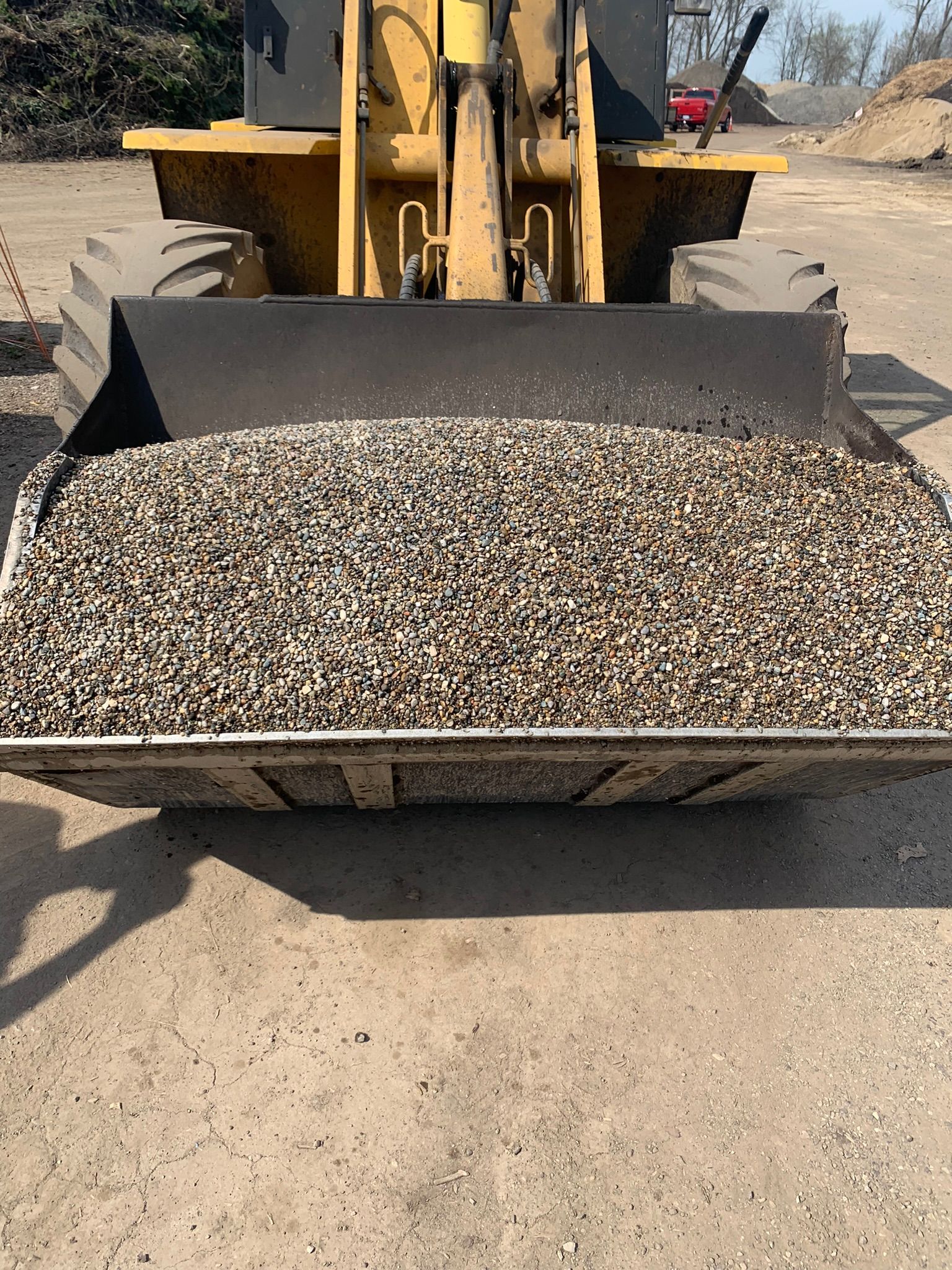 A yellow front-end loader bucket filled with small, dark gravel, parked on a dirt lot.