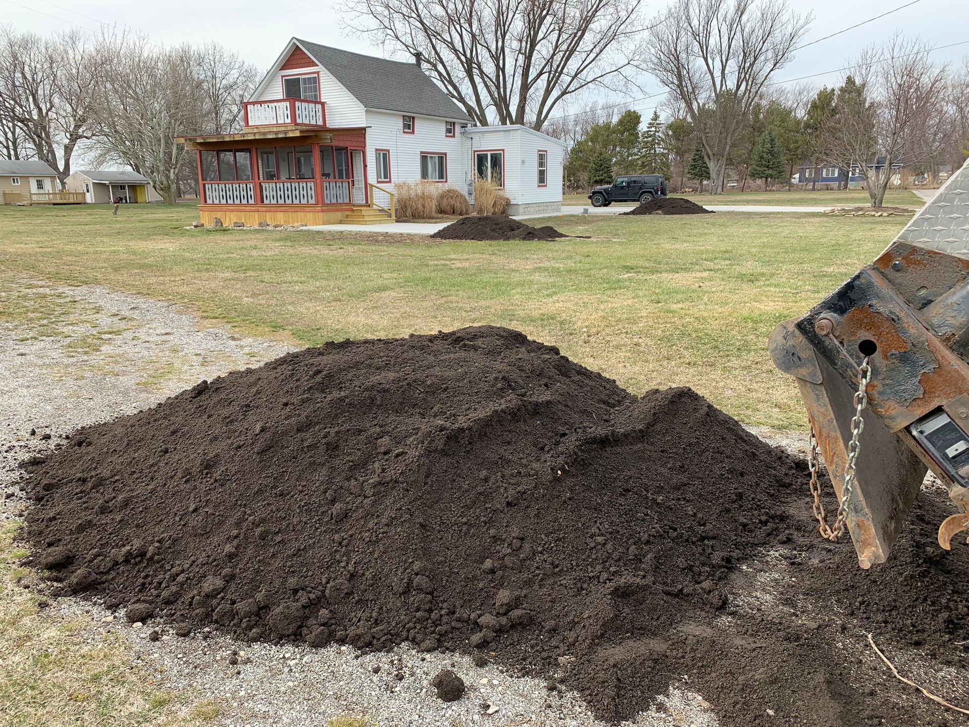 A large pile of dark topsoil sits on a gravel driveway in front of a white house with a porch.