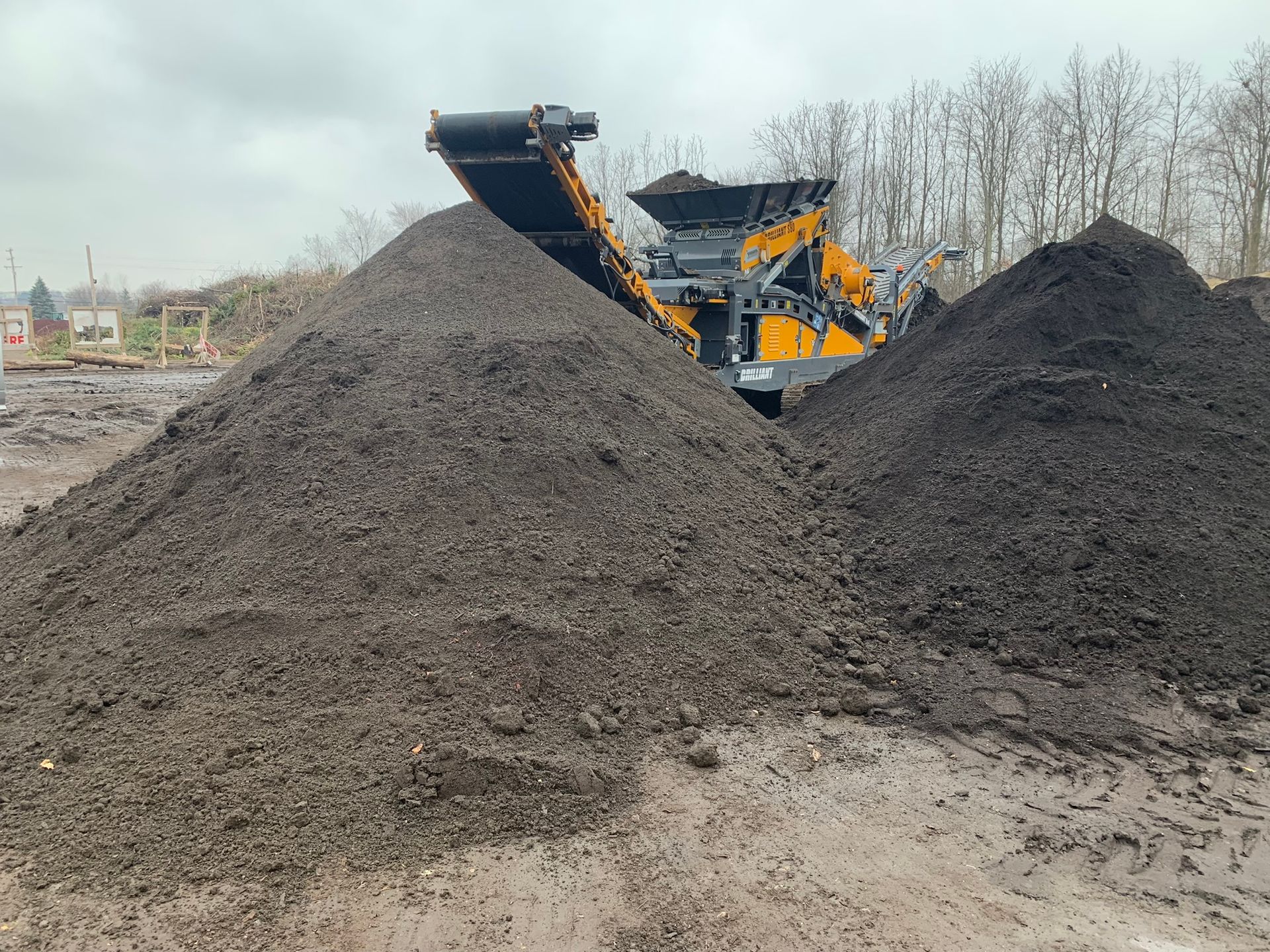 Yellow industrial machinery piles dark soil into large mounds on a cloudy day.