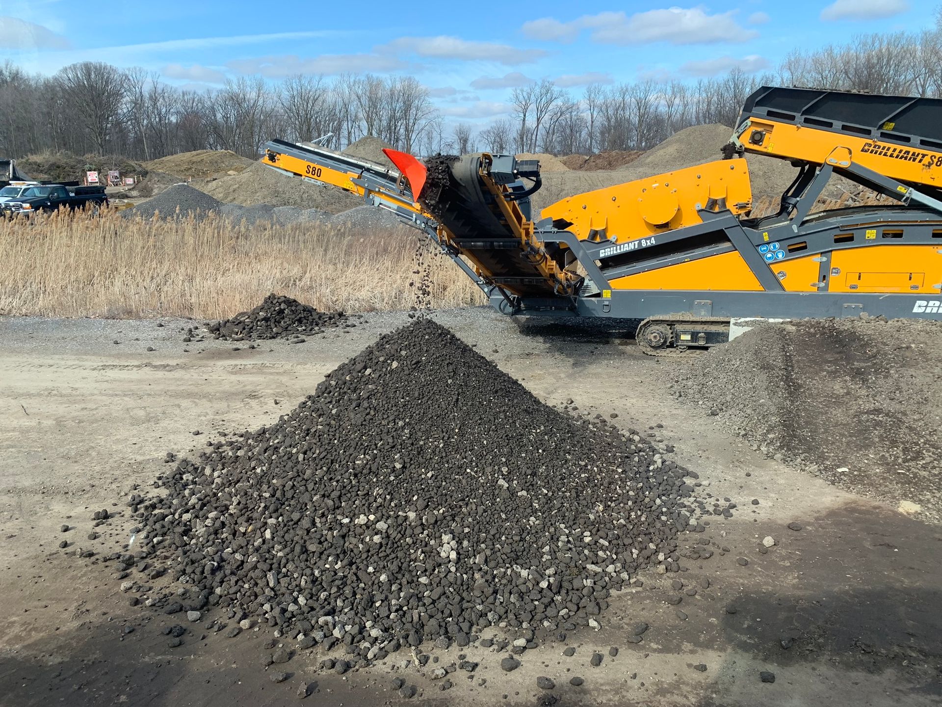 A yellow industrial screening machine discharges a large pile of dark, crushed aggregate material onto a dirt lot.