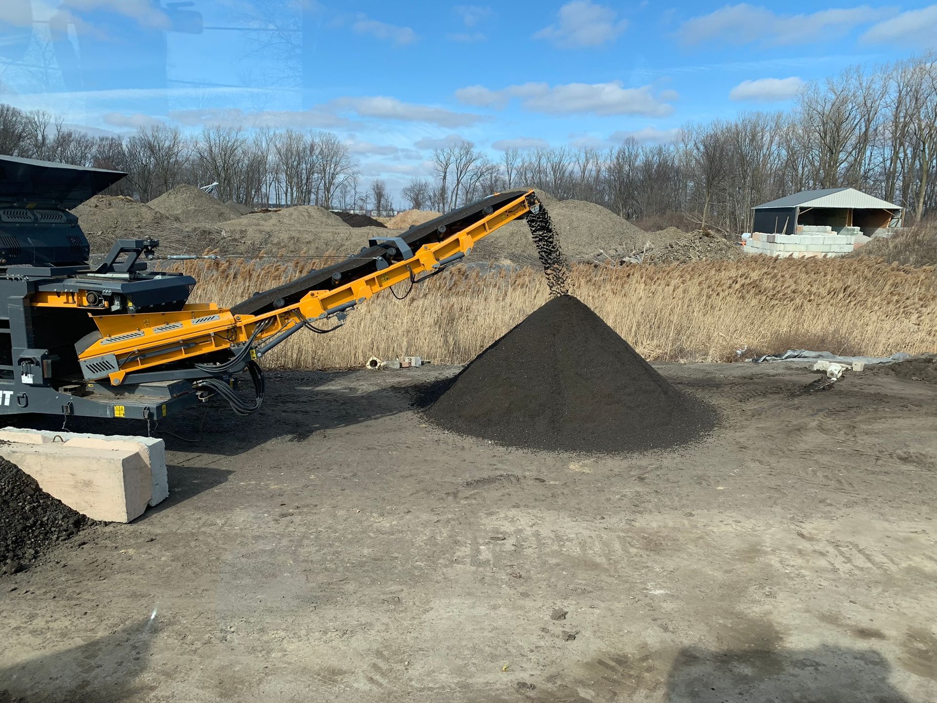 A yellow industrial conveyor machine deposits a dark pile of soil onto the ground under a clear blue sky.