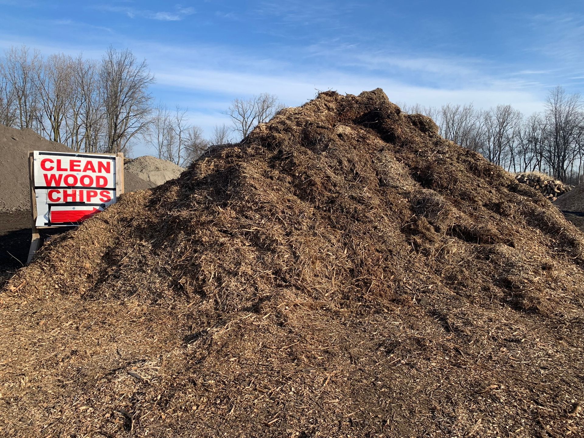 A large, conical pile of brown wood chips against a blue sky, with a sign reading 